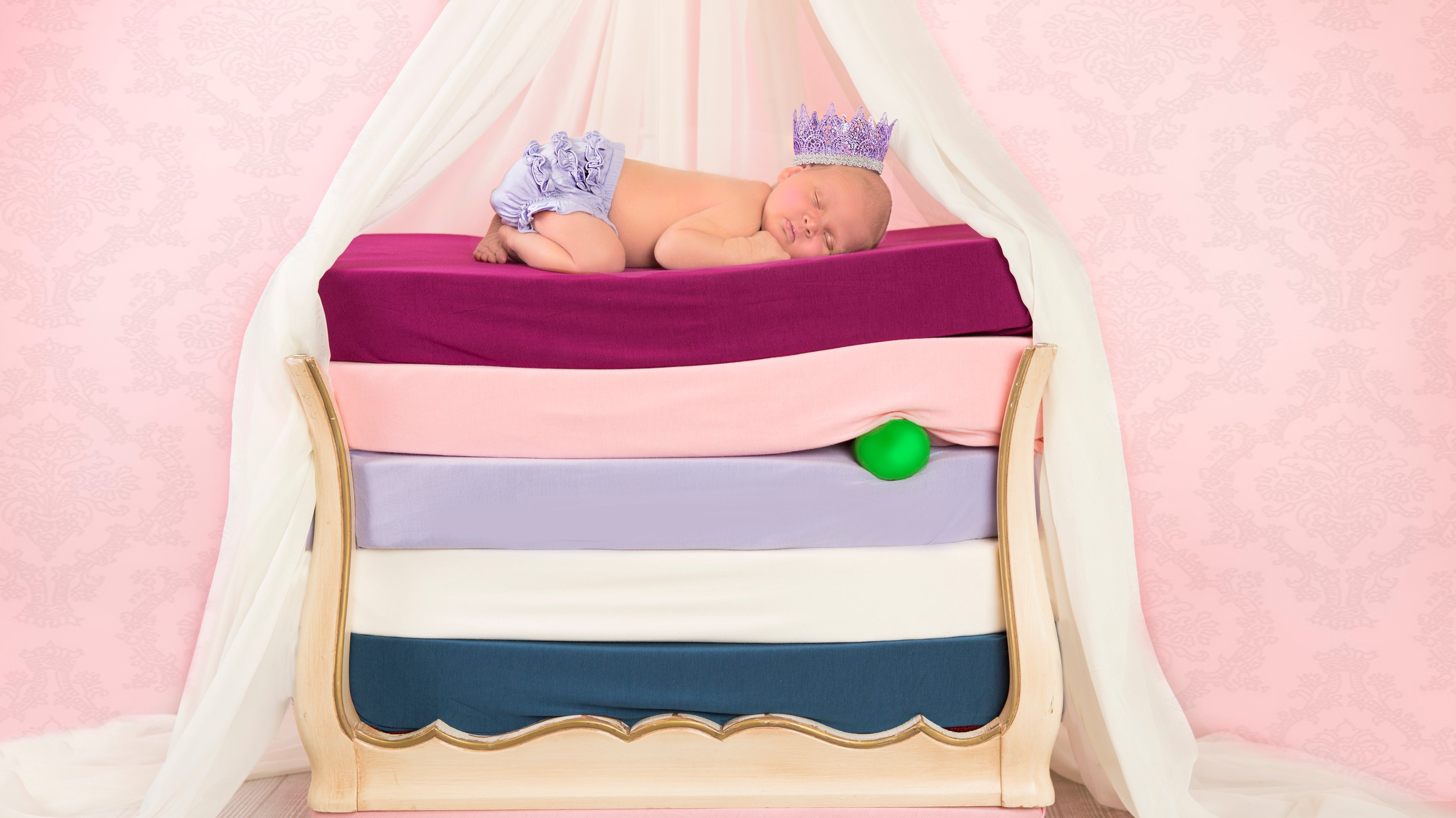 A sleeping baby wearing a crown sleeps on top of a stack of beds with bamboo crib sheets on them. Made to evoke the story of the Princess and the Pea.