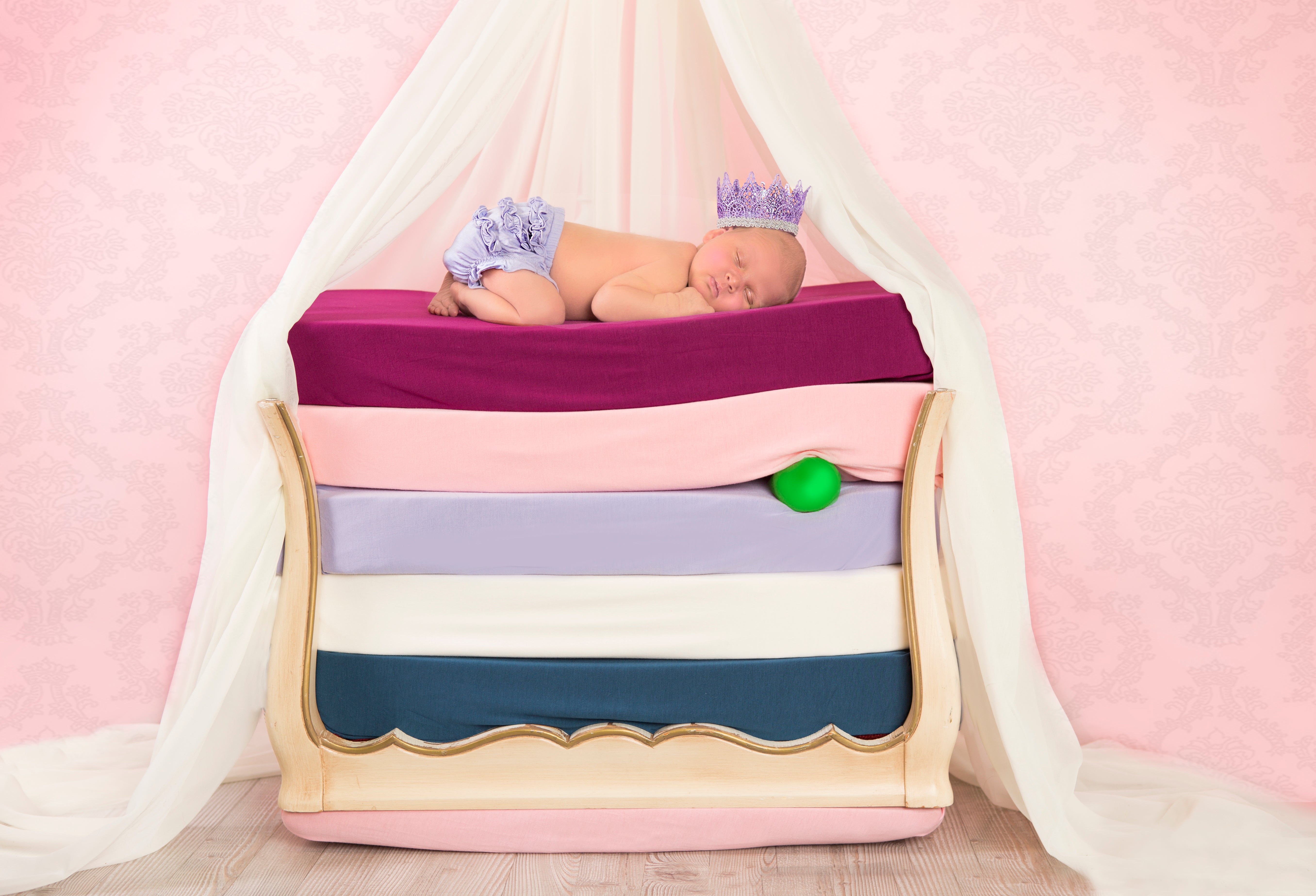 A sleeping baby wearing a crown sleeps on top of a stack of beds with bamboo crib sheets on them. Made to evoke the story of the Princess and the Pea.