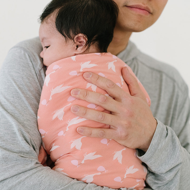ブラッシュのコウノトリ柄レシービングブランケットに包まれ、保護者の胸元に抱かれる赤ちゃん。