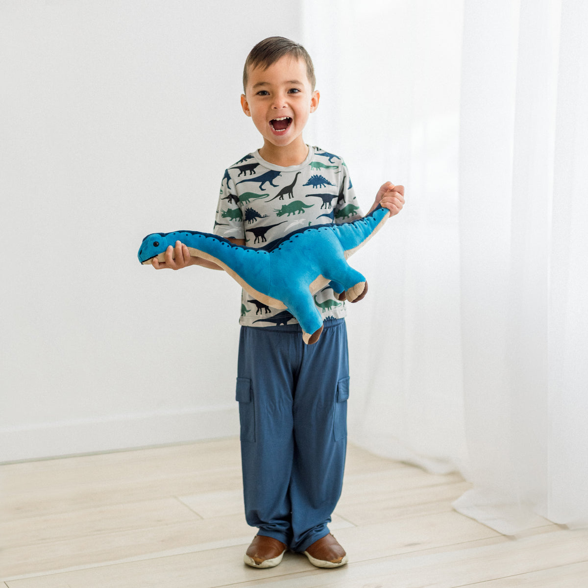 Dove Dino Museumの恐竜柄半袖クルーネックTシャツを着て、青い恐竜のぬいぐるみを持つモデルの全身