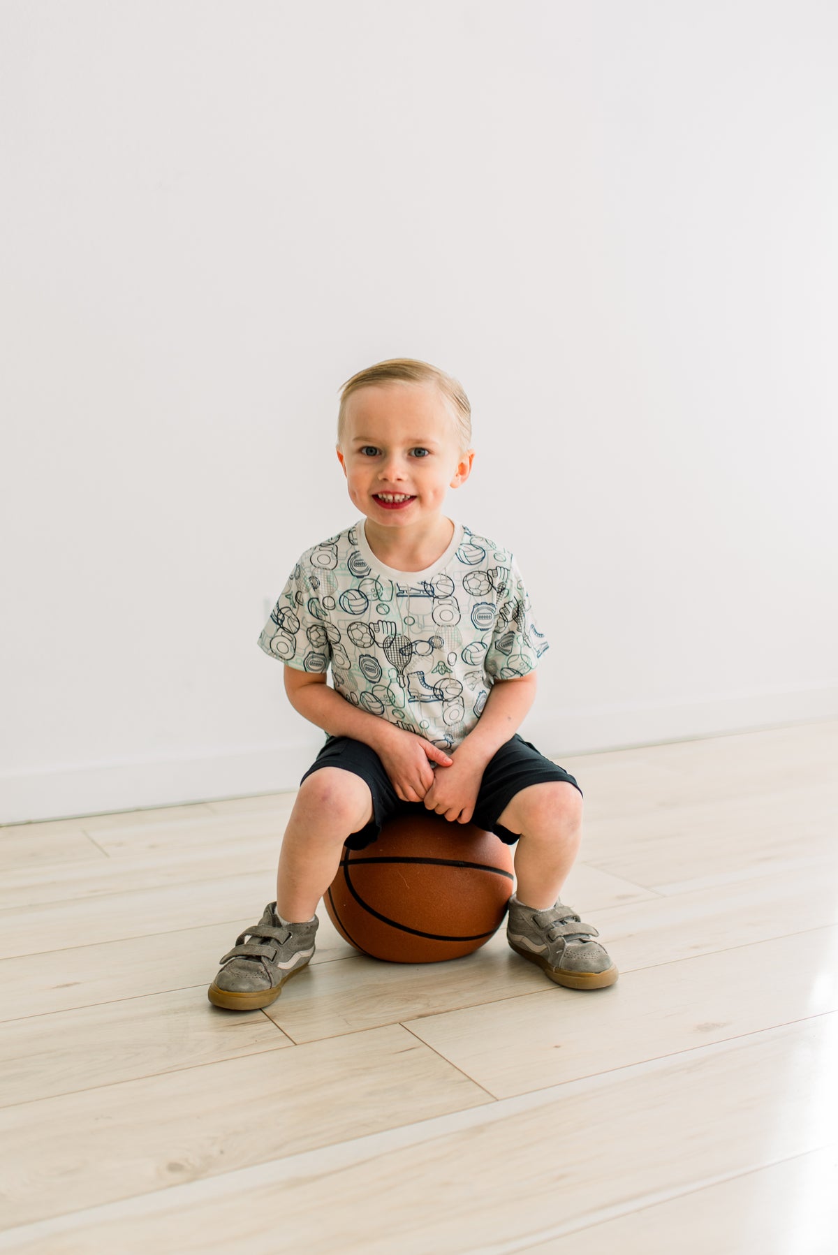 Natural Sports Gear柄の半袖クルーネックTシャツを着て、バスケットボールに座る子どもの正面