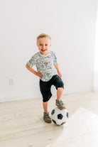 Natural Sports Gear柄の半袖クルーネックTシャツを着て、サッカーボールに片足をのせて立つ子どもの全身
